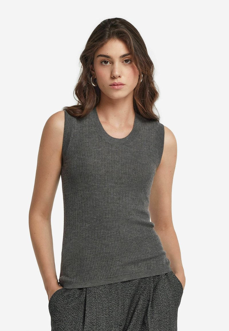 Ung kvinna med axellångt vågigt brunt hår som bär silvriga hoops örhängen och en åtsittande ärmlös ribbad grå topp med texturerade grå byxor.