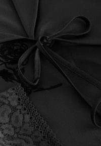 Schwarzes Kleidungsstück aus Stoff mit satinartigem Finish, mit einer Schleife in der Mitte, Spitzenverzierung und einem floralen, in Schwarz geprägten Design.