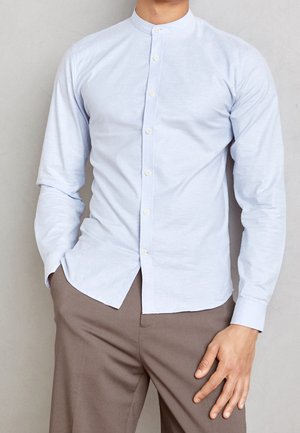 Mann trägt ein hellblaues Hemd mit Stehkragen, die linke Hand in der Tasche und die rechte Hand ruht auf brauner Hose.