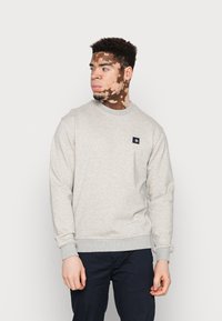 Ung mann med vitiligo iført en lys grå sweatshirt og mørke bukser, som ser til venstre mot en ensfarget hvit bakgrunn.
