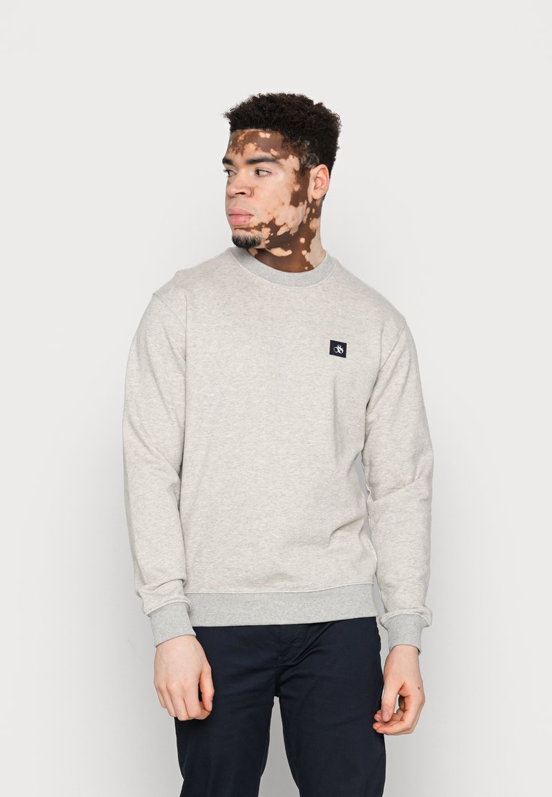 Ung mann med vitiligo iført en lys grå sweatshirt og mørke bukser, som ser til venstre mot en ensfarget hvit bakgrunn.