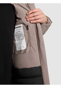 Padded jacket s béžovým vrškem a černou spodní částí. Má zipovou vnitřní kapsu a na vnitřní straně je viditelná etiketa s pokyny k údržbě.