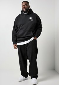 Hombre con sudadera negra con capucha y logo blanco, pantalones negros sueltos y zapatillas blancas, de pie con una mano en el bolsillo frente a un fondo liso.