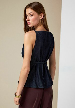 Naine, kellel on pikad pruunid juuksed, kellel on seljal seotav varrukateta tume teksatopp ja bordoopunased püksid, vaatab üle õla.