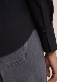 Czarny longsleeve z gładką fakturą, z zaokrąglonym dołem i mankietem zapinanym na guziki, w połączeniu z szarymi dżinsami z widoczną kieszenią z tyłu.
