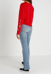 Röd långärmad tröja med blommig broderi på vänster ärm, tillsammans med ljusblå denim flare jeans och svarta sneakers.