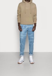 Beige huppari, jossa on kiristysnauhat ja etutasku, yhdistetty vaaleansinisiin farkkuihin. Juoksutyyppiset valkoiset tennarit kumipohjilla.