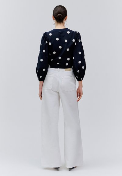 Sötét hajú, kontyba tűzött nő fekete, fehér pöttyös hosszú ujjú blúzt és magas derekú, széles fehér nadrágot visel szürke háttér előtt.