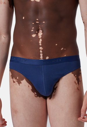 Marineblaue Baumwollslips mit glatter Textur und einem dezenten Bund, der mit dem eingeprägten "SKINY"-Logo versehen ist. Hautfarbe mit Vitiligo-Mustern.