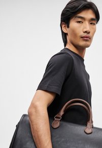 Melnas ādas rokassoma ar brūnām ādas rokturiem, ar gludu tekstūru un minimālistisku dizainu. Rokturiem ir šuvju akcenti.