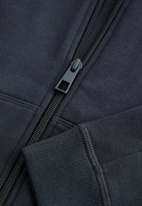 Sweat-shirt noir zippé avec une fermeture éclair robuste, des poignets côtelés et un tissu doux et texturé. Points de couture détaillés le long des coutures.
