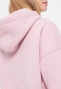 Roosa kapuutsiga sviiter pehme tekstuuri ja vabalt lõikega. Omab nööriga kapuutsi ja ribatekke. Nähtavad õmblusaktsendid.