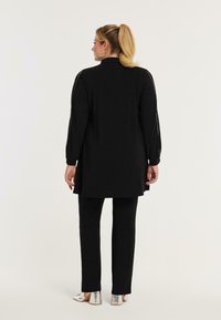 Zwarte lange tuniek met lange mouwen en een rechte snit, gecombineerd met zwarte broek. De outfit heeft een gladde textuur en een minimalistisch ontwerp.