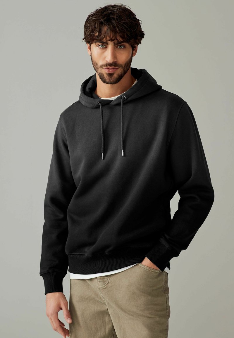 Black hoodie with a drawstring, made of soft cotton blend fabric. Features ribbed cuffs and hem, worn over a white shirt, with a relaxed fit.