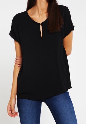 Femme portant un chemisier noir ample à manches courtes avec une petite ouverture sur le devant et une boucle dorée, associé à un jean bleu ajusté.