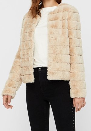 Femme portant une veste en fausse fourrure beige et douce sur un haut blanc et un pantalon noir avec des détails cloutés sur les côtés.