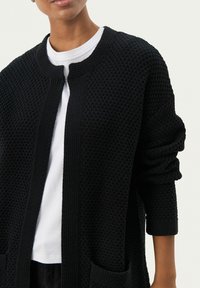 Svart stickad cardigan med en texturerad yta, öppen framtill och två framfickor. Har en rund halsringning och uppvikt trekvartslånga ärmar.