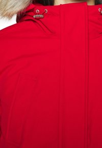 Parka roja con capucha forrada de piel, que presenta herrajes metálicos, un cierre de cremallera y un bolsillo lateral. Tejido suave con un acabado mate.