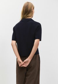 Blå kortærmet sweater med ribbet krave og struktureret strik, båret med brune bukser med striber. Bagsiden viser en afslappet pasform.