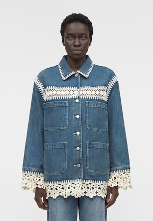 Γυναίκα που φοράει μπλε τζιν μπουφάν με λευκή φλοράλ δαντέλα στο γιακά, το στήθος, τις μανσέτες και το τελείωμα, στέκεται μπροστά σε απλό φόντο.