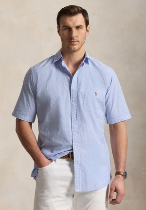 Bărbat în picioare purtând o cămașă cu mâneci scurte, cu dungi albastre și albe, pantaloni albi și o curea maro.