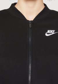 Černá zipová bunda vyrobená z hladké látky, s vysokým límcem a bílým logem Nike na levém prsu blízko zipu.