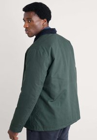 Chaqueta verde con cuello de pana, que presenta un corte relajado y mangas largas. Textura suave y diseño minimalista sin logos visibles.