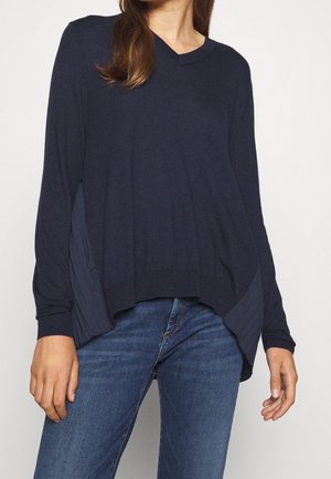 Femme portant un pull ample bleu marine à col en V avec des panneaux tricotés sur les côtés et un jean en denim bleu, debout devant un fond blanc uni.
