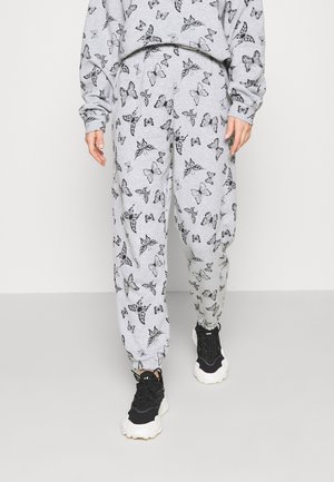 Osoba ubrana w szare spodnie dresowe i pasującą bluzę z czarnymi nadrukami motyli, w połączeniu z czarnymi tenisówkami z białymi podeszwami.