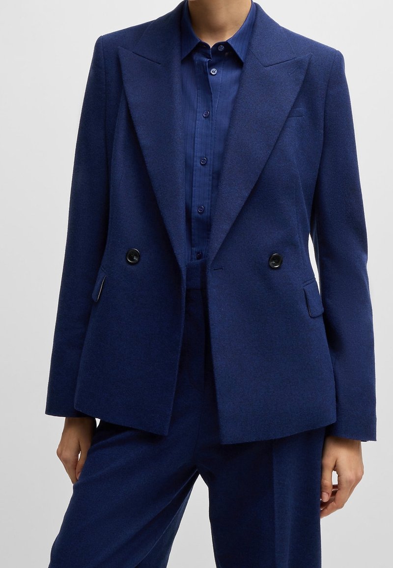 Personne portant un blazer bleu marine cintré croisé avec un pantalon assorti et une chemise bleue boutonnée.