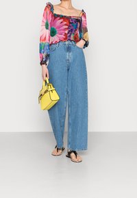 Bunt gemustertes Crop-Top mit langen Ärmeln, kombiniert mit hochgeschnittenen blauen Denim-Breite-Hosen und einer kleinen gelben Handtasche.