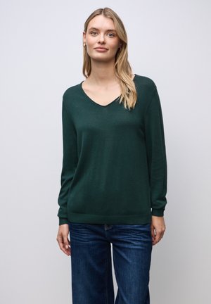 Pull en tricot léger vert foncé avec un col en V, des manches longues et des poignets et un ourlet côtelés. Porte avec un jean bleu à jambes larges.