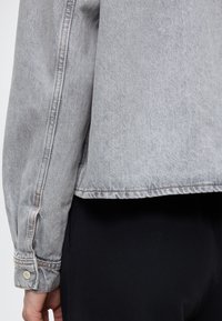 Szürke farmerdzseki enyhén rövidített szabással, varrott varrásokkal és gombos mandzsetta részlettel. Az anyag könnyű, sima tapintású.