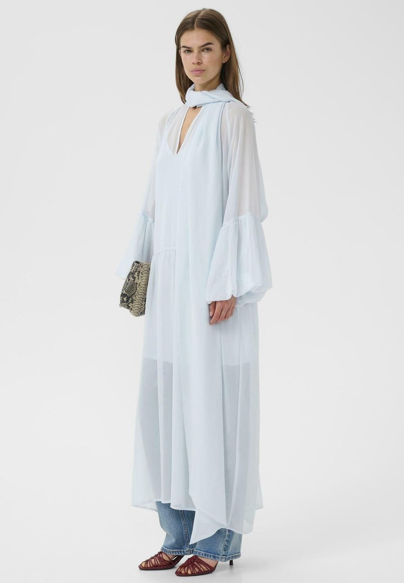Femme portant une robe légère bleu clair transparente avec des manches ballon sur un jean, tenant une pochette à imprimé serpent, debout sur un fond blanc.