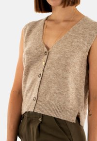 Cardigan senza maniche beige, realizzato in tessuto morbido, con scollatura a V, bottoni frontali e orlo a costine. Presenta fessure laterali per un comfort aggiuntivo.