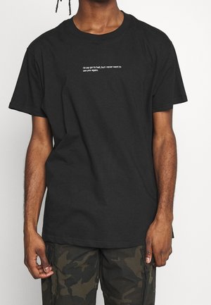 Personne portant un t-shirt noir avec un texte blanc indiquant "I'd say go to hell, but I never want to see you again.". Pantalon camouflage visible.