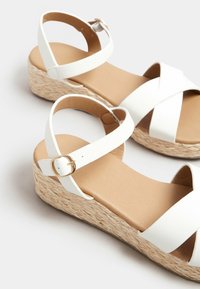 Sandali con zeppa bianchi, con cinturini incrociati, chiusura con fibbia e una piattaforma testurizzata simile alla paglia. Soletta liscia di colore beige.