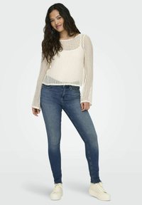 Suéter de punto color crema con mangas largas y un patrón texturizado, combinado con jeans ajustados azules y zapatillas blancas.