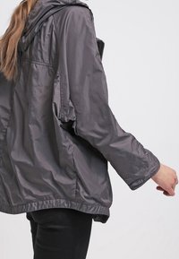 Grå nylonjakke med hætte, løs pasform og lange ærmer. Har en glat tekstur og elastisk kant, vist fra en sidevinkel.