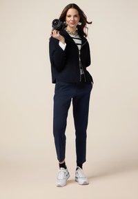 Marinfärgad zip-up jacka över en randig topp, ihop med marinfärgade byxor och vita sneakers. Tillbehören inkluderar ett kraftigt halsband och en kamera.