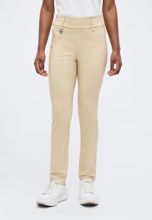 Donna che indossa pantaloni beige aderenti e sneakers bianchi, in piedi contro uno sfondo bianco uniforme.