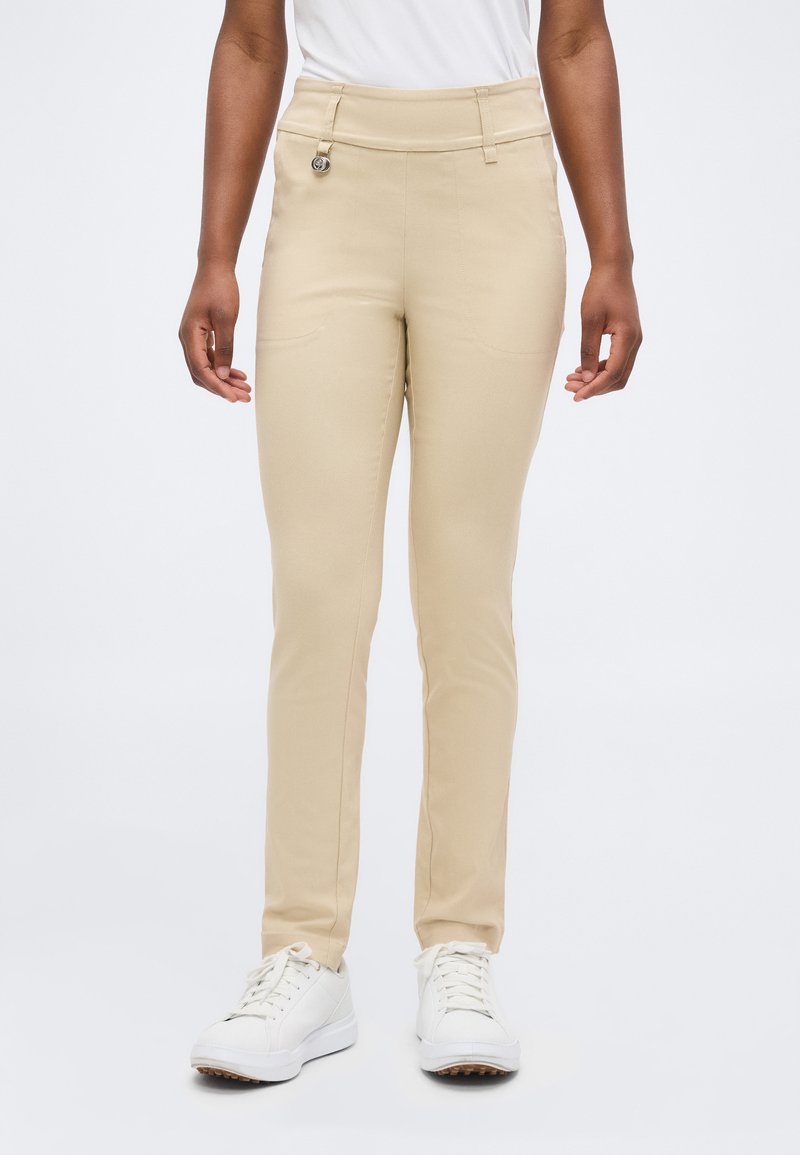 Γυναίκα που φοράει μπεζ παντελόνια slim-fit και λευκά αθλητικά παπούτσια, στέκεται μπροστά σε ένα απλό λευκό φόντο.