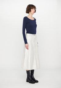 Top a maniche lunghe blu navy abbinato a una lunga gonna di denim bianca. La modella indossa stivali alla caviglia neri e lucidi. Design minimalista con silhouette aderente.