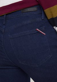 Vaqueros de mezclilla oscuro con un bolsillo trasero que presenta un acento de franjas rojas, blancas y azules y una textura sutil. La etiqueta dice "Tommy Hilfiger."