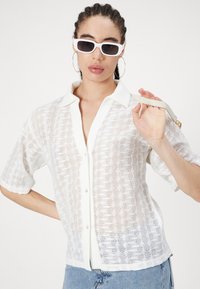Maglietta bianca in pizzo con maniche corte, chiusura frontale a bottoni e un motivo texture. Il modello tiene una tracolla, indossa occhiali da sole bianchi e orecchini a cerchio.