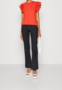 Czerwony krótki top z krótkim rękawem i koronkowymi akcentami, w połączeniu z ciemnymi dzwonowatymi dżinsami i białymi sandałami na platformie. Modelka trzyma małą białą torebkę.