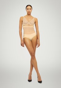 Nude mesh underkläder set bestående av en bralette med dekorativa remmar och matchande högmidjade trosor. Slät textur och minimalistisk design.