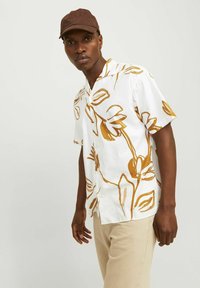 Camicia bianca a maniche corte con stampa floreale marrone, tessuto leggero, colletto a punte e bottoni. Indossata con pantaloni di colore chiaro.