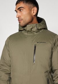 Olivgrüne, mit Kapuze versehene Jacke mit einem Reißverschluss-Brusttasche, strukturiertem Stoff und dem Columbia-Logo auf der linken Seite.