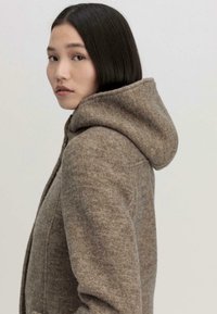 Bruin wollen jas met een capuchon; heeft een zachte, gestructureerde afwerking en een getailleerd ontwerp, met stiksels en naden langs de zijkant.